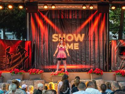 Mädchen in rosa Kleid singt auf Bühne vor Publikum mit roter Beleuchtung und Blumen.