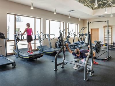 Fitnessraum mit Laufbändern, Hantelbank und kräftigen Geräten, helle Wände und große Fenster.