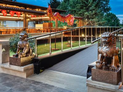 Zwei bronzene Löwenstatuen bewachen einen Parkzugang mit Holzbrücke und chinesischer Architektur.