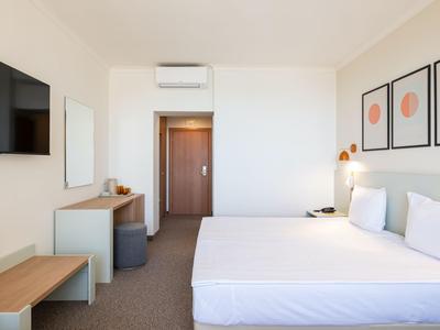 Helles Hotelzimmer mit Doppelbett, TV, Hocker und minimalistischem Design.