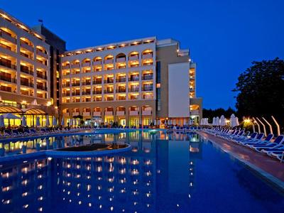 Ein beleuchtetes Hotelgebäude mit vielen Balkonen, angrenzend an einen großen Pool bei Abenddämmerung.