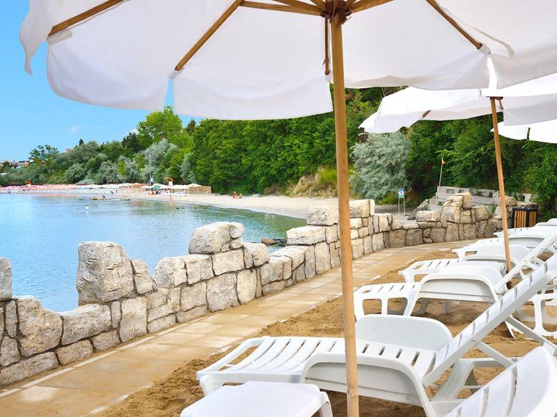 Lettini bianchi e ombrelloni lungo una passeggiata con muro di pietra vicino all'acqua.