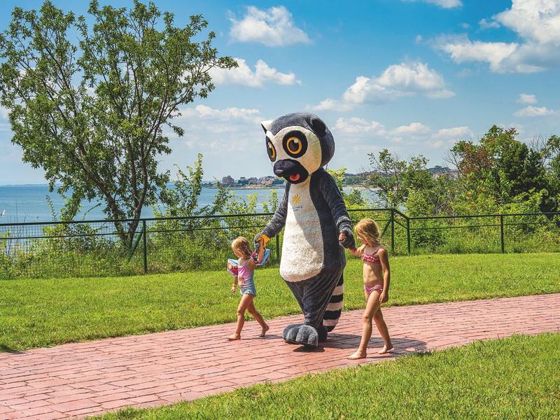 Due bambini camminano accanto a una persona in costume da pinguino su un sentiero lastricato in un parco con un lago sullo sfondo.