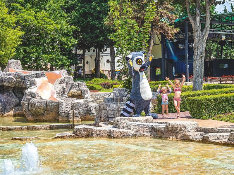 Un parco a tema con una cascata, una grande mascotte animale e bambini che giocano vicino all'acqua.