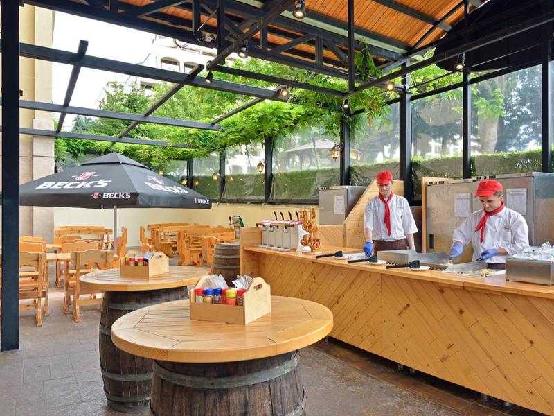 Area esterna di un caffè con bancone in legno, tavoli rotondi e due dipendenti.