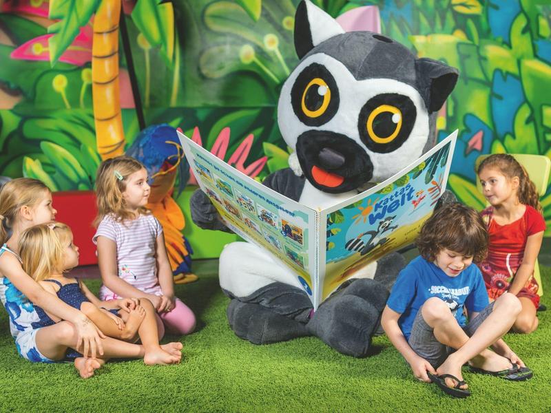 Bambini seduti su pavimento verde ascoltano una mascotte con testa di animale che legge un libro.