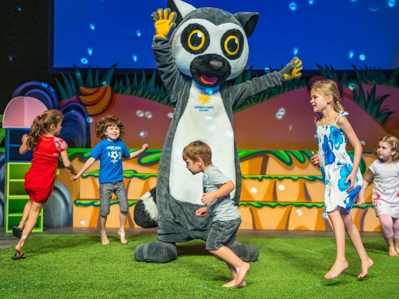 Bambini che giocano e ballano con una grande mascotte animale su uno sfondo di scena colorato.