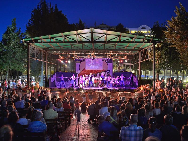 Concerto serale all'aperto con grande pubblico e palco illuminato in un parco.