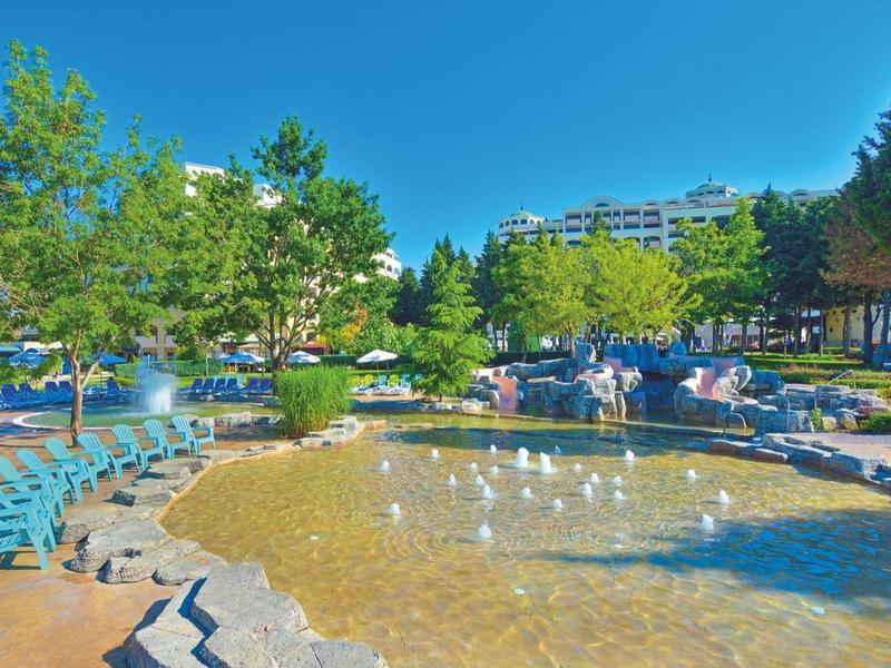 Grande piscina all'aperto con lettini, alberi e edifici sullo sfondo sotto un cielo sereno.