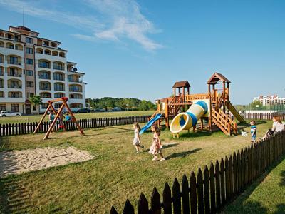Spielplatz mit Rutschen und Sandkasten neben einem Hotelgebäude bei klarem Himmel.