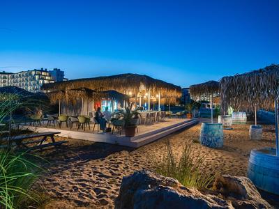 Strandbar mit Strohdächern und Sitzgelegenheiten am Sand unter blauem Abendhimmel.