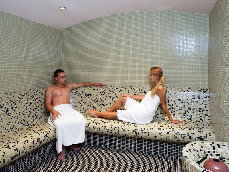 A man and woman sitting on mosaic benches in a sauna room, both wrapped in white towels.