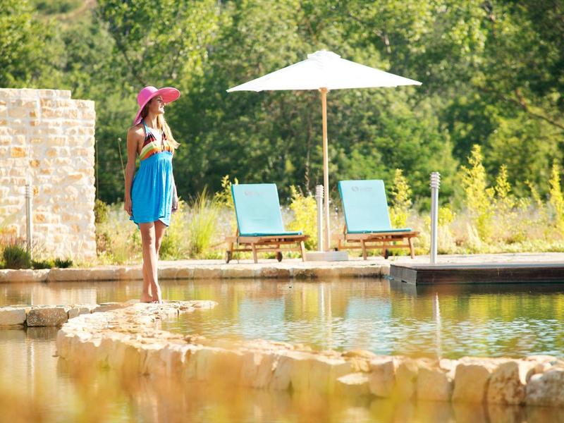 Woman in blue dress and red hat by pool with lounge chairs and umbrella in green setting