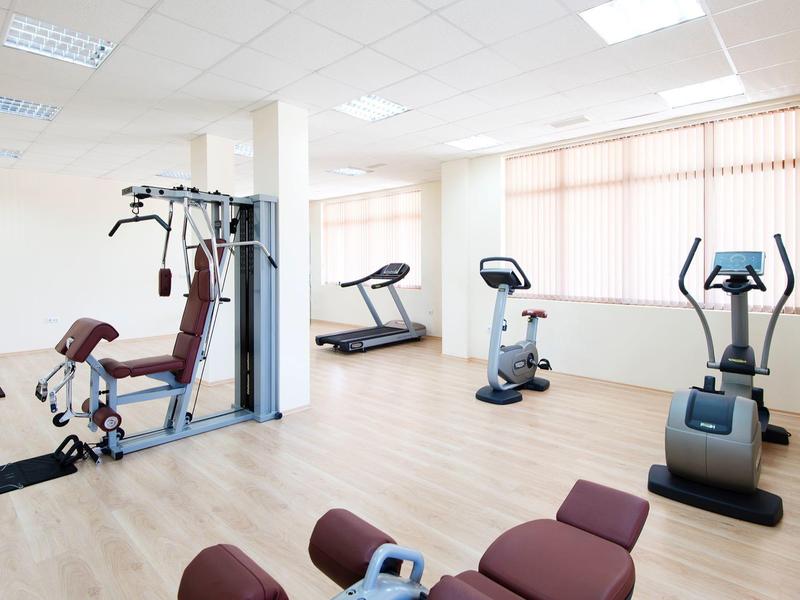 Bright gym room with various exercise machines and large windows with blinds.