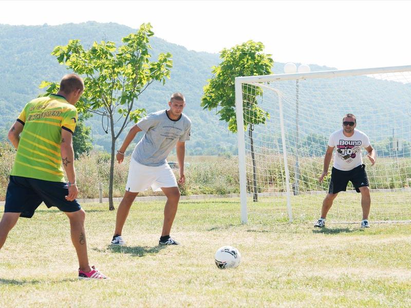 Three men playing soccer on a grassy field in front of a goal with mountains in the background.