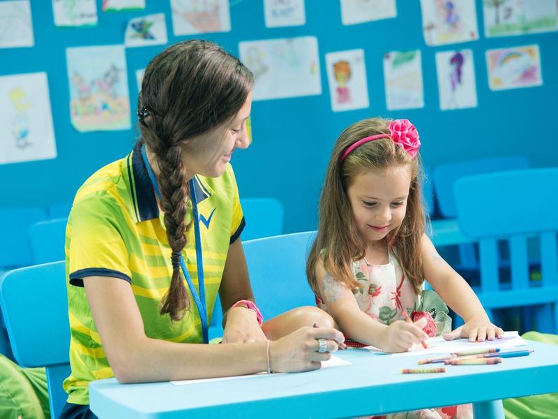 A caregiver helping a girl with drawing at a blue table in a room with children's artwork.