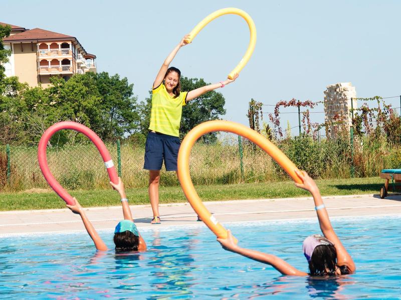 Three children play with colorful pool noodles in a sunny outdoor swimming pool.