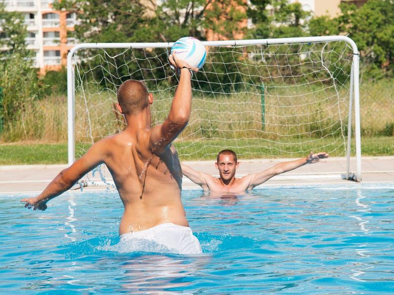 Two men playing water polo in an outdoor pool in front of a soccer goal.