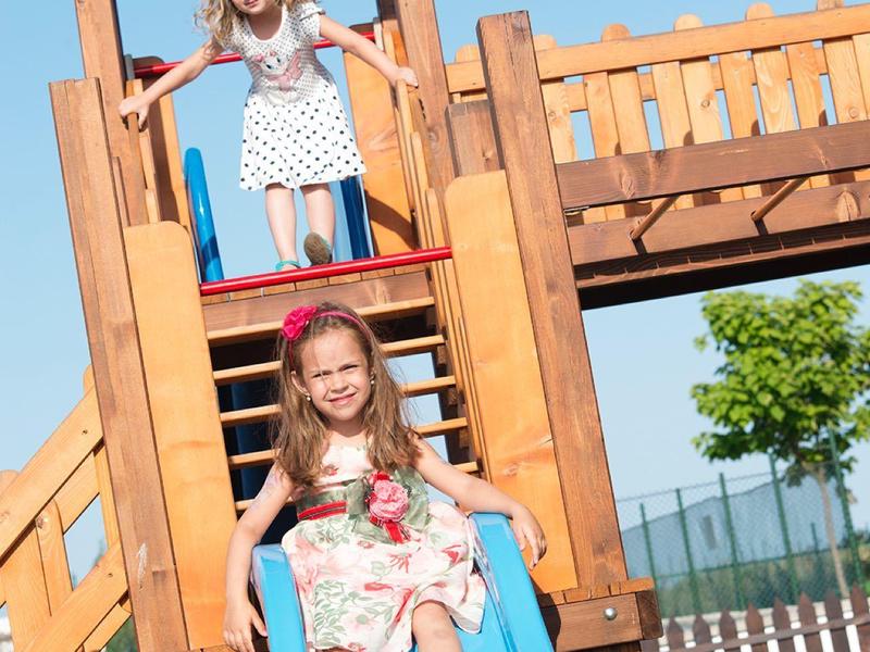 Children playing on a blue slide in a wooden playground structure under clear sky.