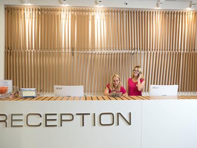 Dvě ženy u bílé recepce s dřevěnou texturovanou stěnou v pozadí.