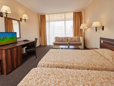 Hotelový pokoj se dvěma postelemi, pohovkou, psacím stolem a televizí u velkého okna se závěsy.