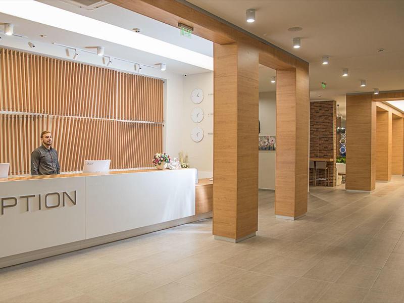 Moderní hotelová recepce s bílým pultem, dřevěnými panely a prostornou chodbou.