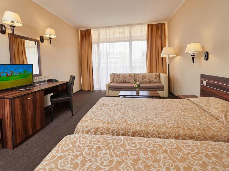 Hotelový pokoj se dvěma postelemi, pohovkou, psacím stolem a televizí u velkého okna se závěsy.