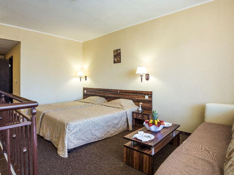 Pohodlný hotelový pokoj s manželskou postelí, malým dřevěným stolem, miskou ovoce a béžovou pohovkou.