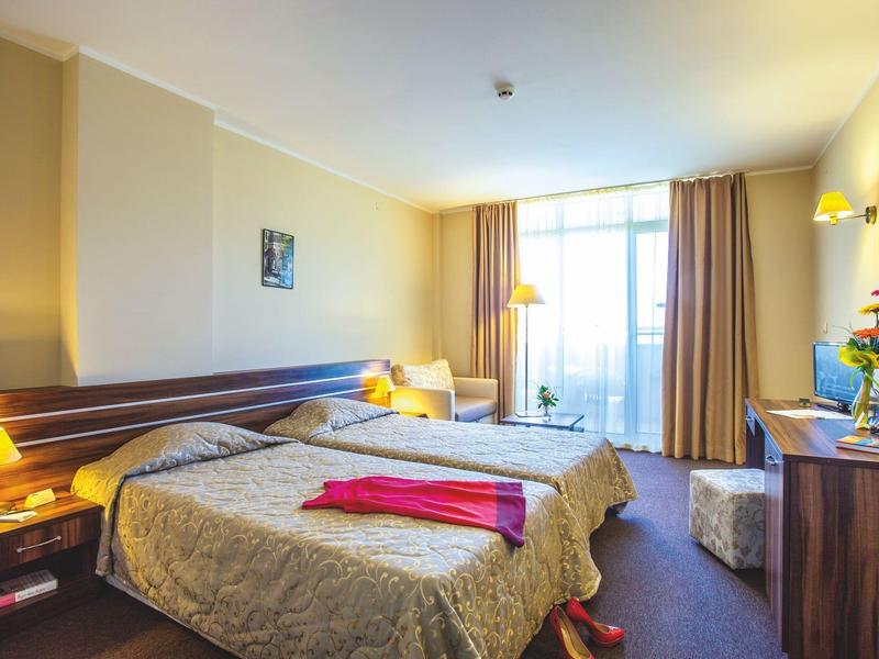 Světý hotelový pokoj s manželskou postelí, křeslem, psacím stolem a velkým oknem s závěsy.