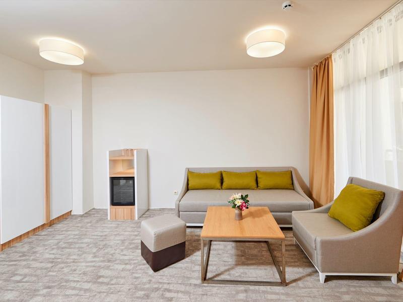 Světý hotelový pokoj se dvěma šedými pohovkami, konferenčním stolkem a velkými okny se závěsy.