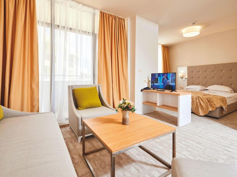 Světý hotelový pokoj s pohovkou, křeslem, dřevěným stolem a postelí vedle televize.