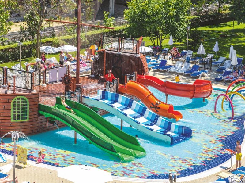 Vodní park s barevnými skluzavkami a lehátky na slunné zelené ploše.