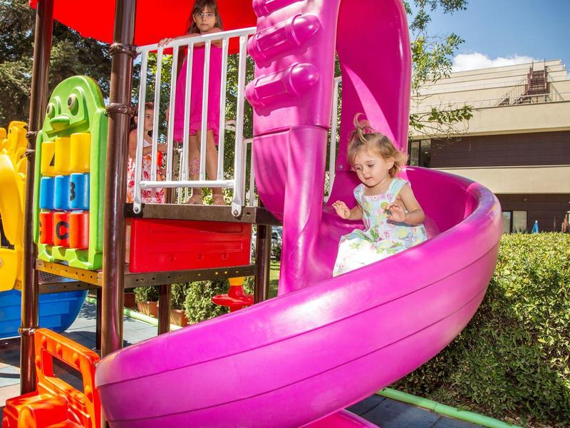Dítě sjíždí jasně růžovou spirálovou skluzavku na barevném hřišti za slunečného dne.