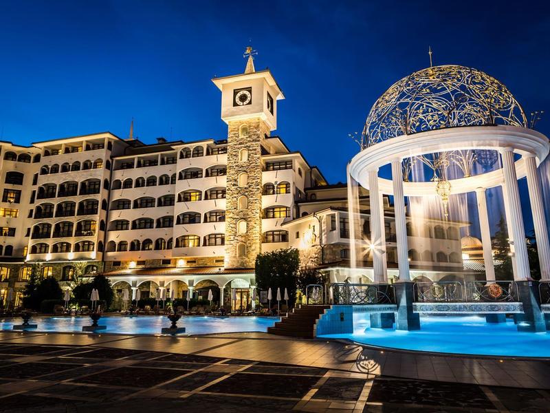 Beleuchtetes Nachtbild eines großen Hotels mit Uhrenturm und blauem Pavillon vor dunklem Himmel.
