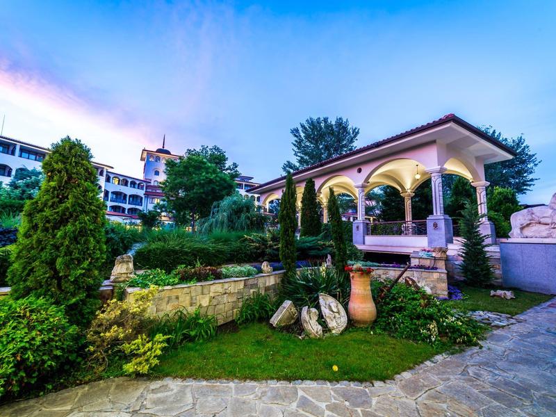 Garten mit Steinen, Zypressen und überdachtem Pavillon unter blauem Himmel bei Sonnenuntergang.