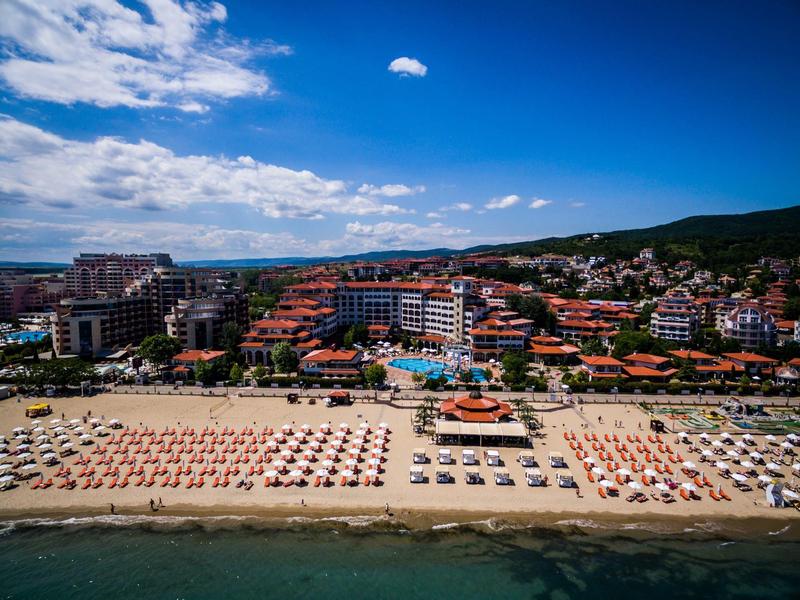 Strand mit vielen Sonnenschirmen und Liegen, dahinter Hotels und bewaldete Hügel unter blauem Himmel.