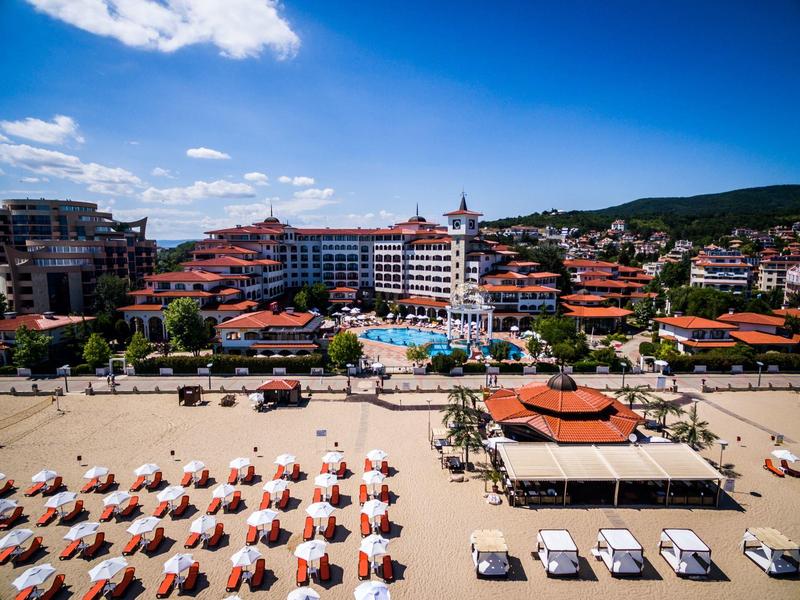 Strand mit vielen weißen Sonnenschirmen, Liegen und roten Dächern vor blauen Himmel und Hügeln.