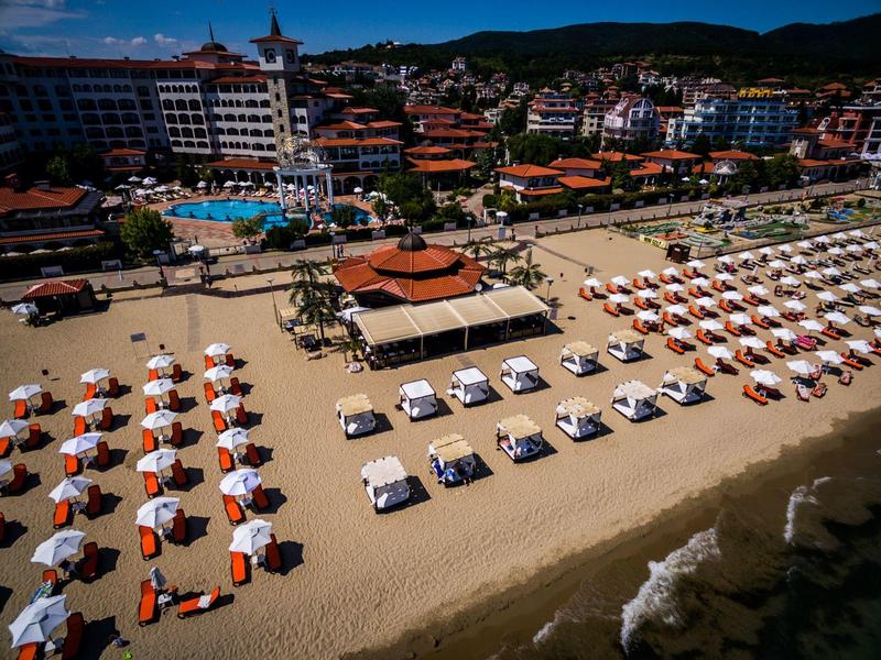 Breiter Sandstrand mit Sonnenschirmen, Liegen und Badehäuschen vor Hotelanlagen und bewaldeten Hügeln.