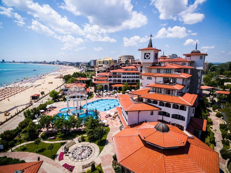 Strandresort mit Pool, mediterranen Gebäuden und Sandstrand unter blauem Himmel mit Wolken.