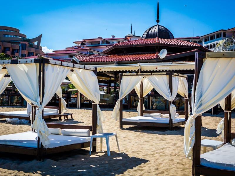 Strandliegen mit weißen Vorhängen auf Sand, im Hintergrund bunte Gebäude unter blauem Himmel.