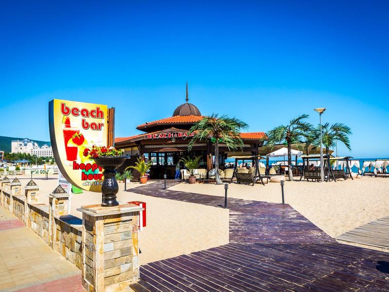 Strandbar mit Sonnenschirmen, Palmen und blauem Himmel in einer sonnigen Küstenanlage.