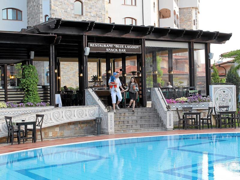 Restaurant mit Terrasse und Tischen neben einem klaren blauen Pool, umgeben von Gebäuden.