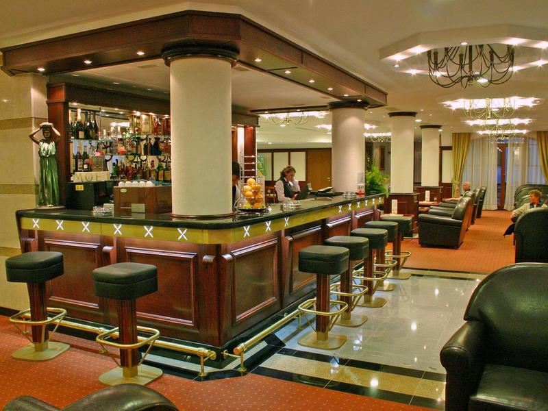 Moderner Barbereich mit Holztheke, grünen Barhockern und Teppichboden in warmen Farben.