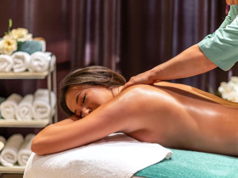 Frau liegt entspannt auf Massagebank, bekommt Rückenmassage, Umgebung mit weißen Handtüchern und Blumen.