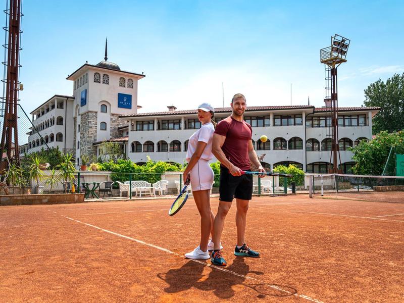 Zwei Tennisspieler auf rotem Sandplatz vor altem, weißem Gebäude mit blauen Fenstern.