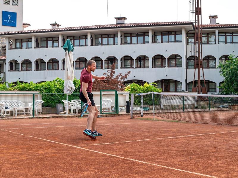 Mann in rotem Shirt spielt Tennis auf rotem Sandplatz vor weißem Gebäude mit Bögen.