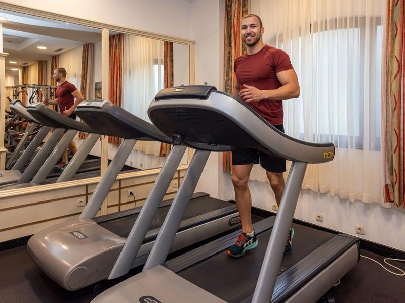 Mann trainiert auf Laufband in kleinem, hellen Raum mit Teppich und Vorhängen.