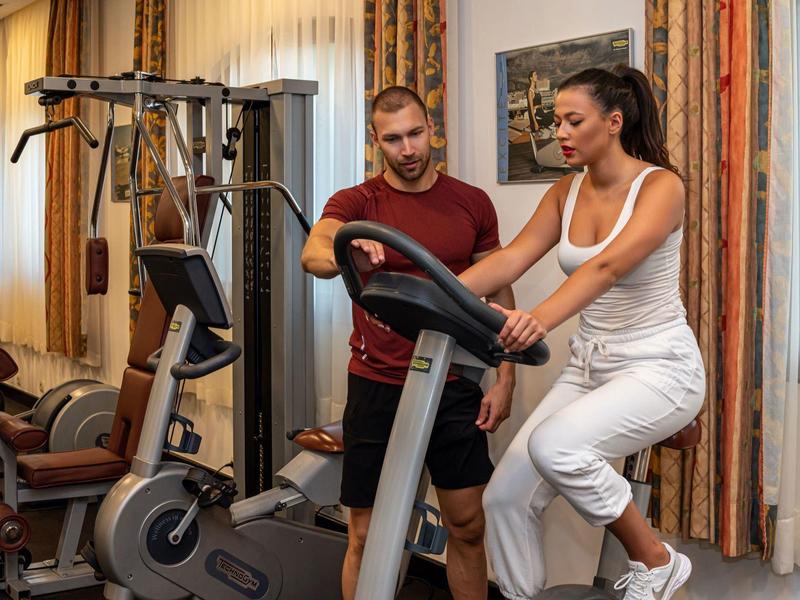 Frau trainiert auf Heimtrainer, Trainer zeigt Anleitung, Raum mit Gardinen und Trainingsgeräten.