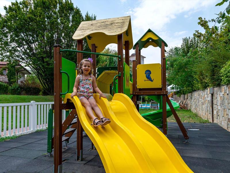 Kind sitzt auf einer gelben Rutsche eines bunten Spielgerüsts im Garten an einem sonnigen Tag.