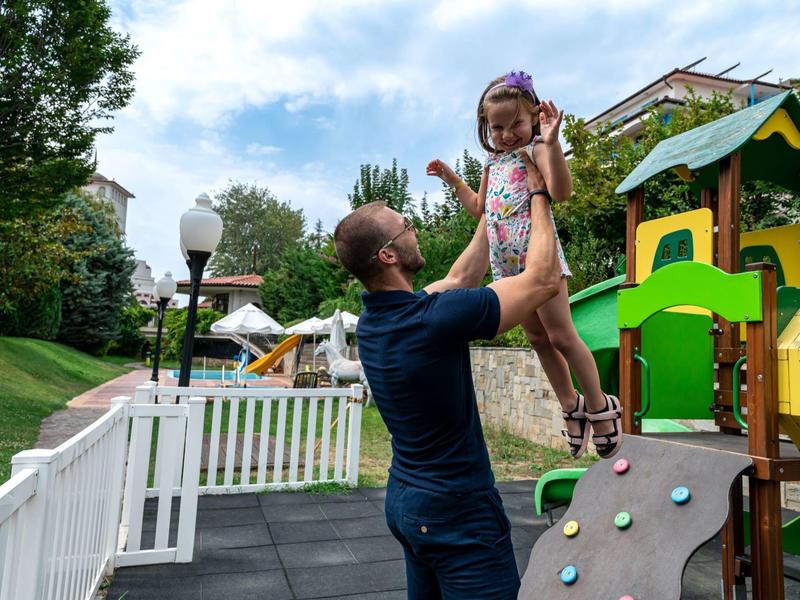 Mann hebt Mädchen spielerisch auf Spielplatz, umgeben von grünem Gras und bunten Spielgeräten.